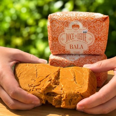 Bala de doce de leite Dona Lázara sendo aberta, com fundo desfocado com plantas verdes.